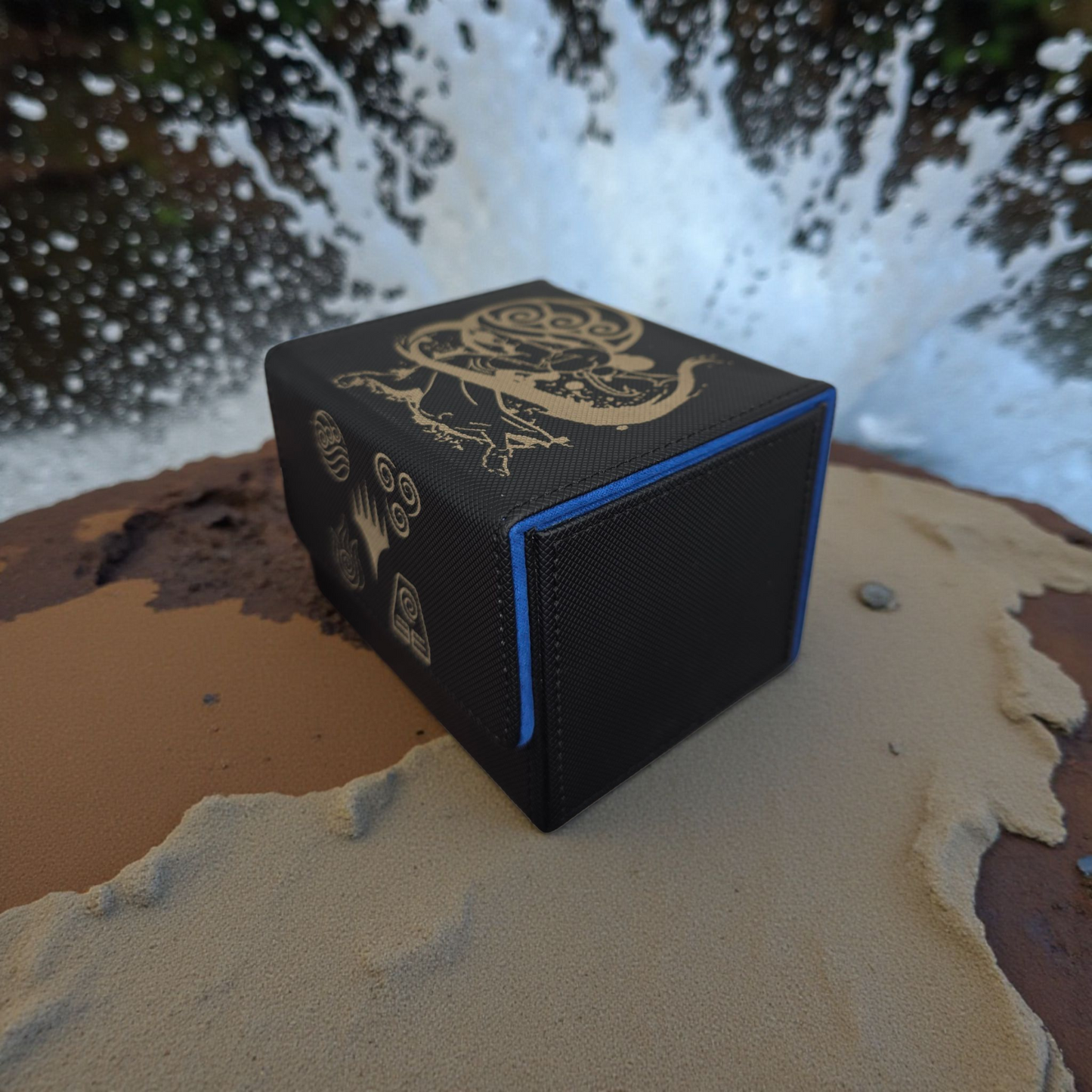 A black and gold folding box with a dragon design on it, placed on a sandy surface with a blurred background of water.