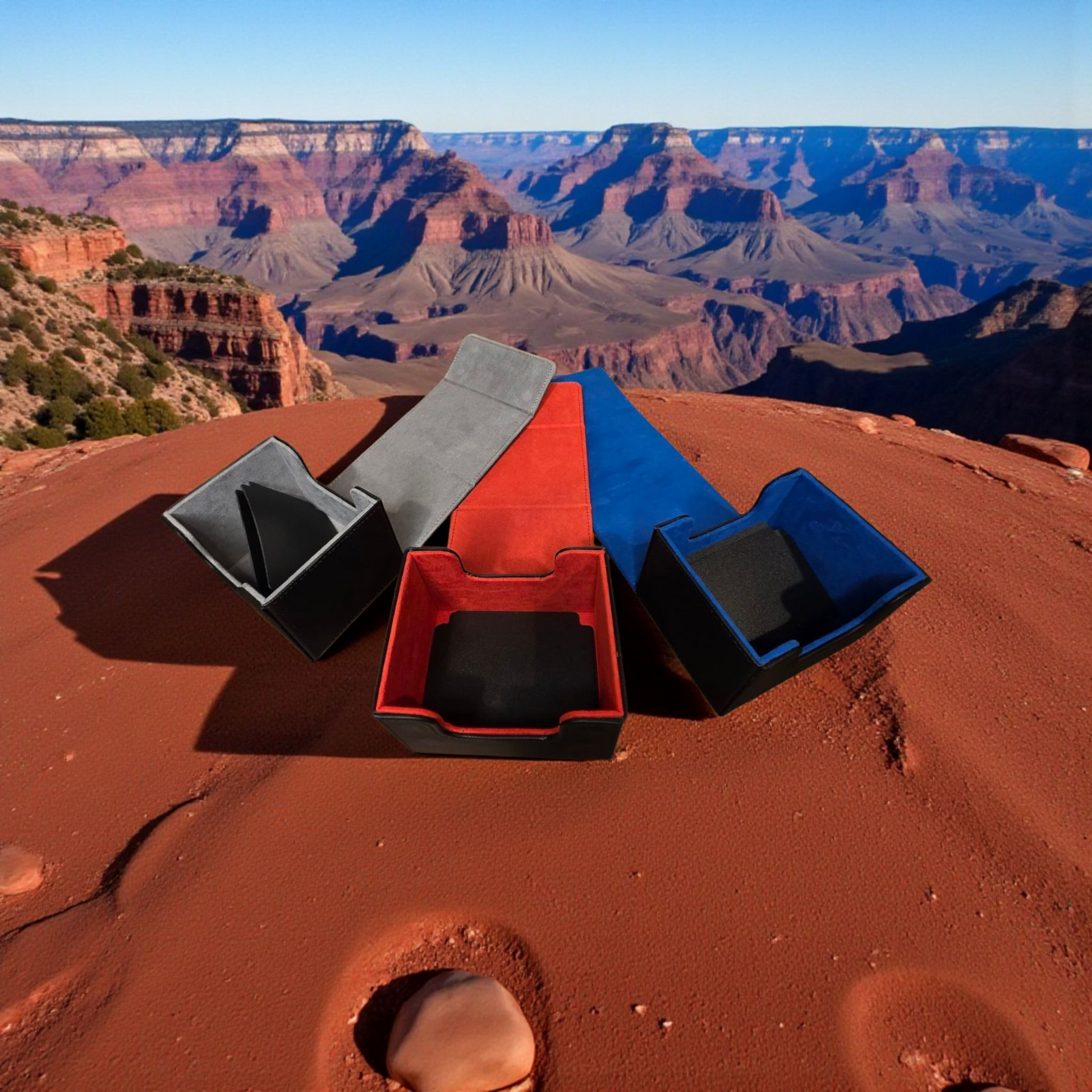 a group of three storage containers, each with a different color: gray, red, and blue. The containers are placed on a red sand surface, with a breathtaking view of the Grand Canyon in the background.